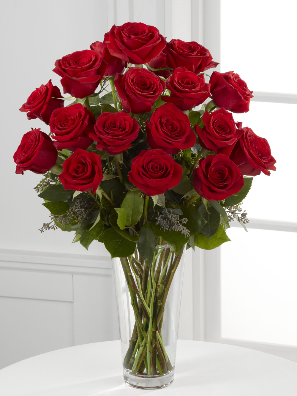 Eighteen red roses with greenery arranged in a tall clear glass vase