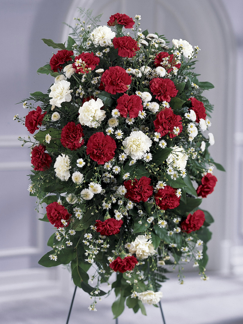 Red and white carnation standing spray with greenery displayed on an easel